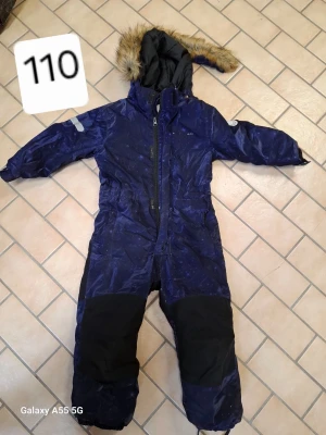 Mörkblå täckbyxa med huva och päls - Täckbyxa från Nordbjørn i mörkblått med svarta förstärkningar på knän och bak. Utrustad med huva och fuskpäls, dragkedja framtill och reflexdetaljer på ärmarna. Perfekt för kalla dagar och snölek.