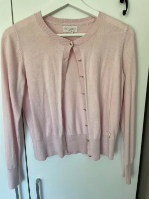 Ljusrosa kofta från Soft Goat - Supersöt ljusrosa kofta från Soft Goat i mjuk och tunn stickning. Rund halsringning, långa ärmar och små guldfärgade knappar framtill. Ribbstickade muddar vid ärmslut och nederkant. Perfekt för lager på lager och en clean look. Liten fläck, men tror det är cola