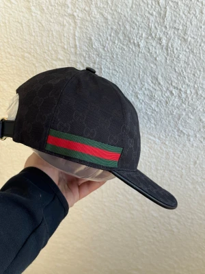 Svart keps från Gucci med GG-mönster - Snygg svart keps från Gucci med klassiskt GG-mönster över hela kepsen. På sidan finns den ikoniska gröna och röda randen. Justerbar rem baktill i läder och böjd skärm. Perfekt för dig som vill ha en stilren och lyxig accessoar.