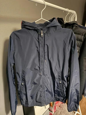 Mörkblå vindjacka från Moncler - Snygg mörkblå vindjacka från Moncler med huva och dragkedja framtill. Jackan har två fickor med tryckknappar och en vit-röd rand på huvan. Moncler-logga på ärmen. Tillverkad i lätt, glansigt material som står emot vind.
