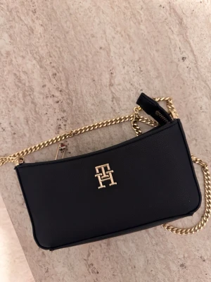 Svart axelväska Tommy Hilfiger - Snygg svart axelväska från Tommy Hilfiger med guldfärgad kedjerem och TH-logga framtill. Väskan har en stilren, rektangulär form och är tillverkad i slätt konstläder. I fint skick!