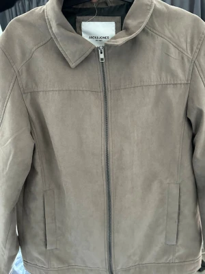 Beige mockajacka från Jack & Jones - Stilren beige mockajacka från Jack & Jones med rak passform och klassisk krage. Jackan har dragkedja framtill och två diskreta sidofickor. Perfekt för dig som gillar en enkel men snygg look.
