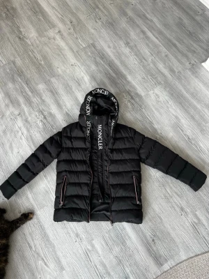 Moncler jacka - Snygg svart dunjacka från Moncler med huva och logotyp på ärmen. Jackan har dragkedja framtill, två fickor med dragkedja och Moncler-tryck på insidan av huvan. Perfekt för kalla vinterdagar och riktigt trendig look. Den passar mig som är 1,60