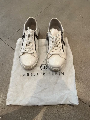 Vita Philipp Plein sneakers med dragkedja - Snygga vita sneakers från Philipp Plein med croco-mönstrad yta och coola silverfärgade dragkedjor på sidorna. Klassisk låg modell med vita snören och detaljer i metall. Perfekta för dig som vill sticka ut med exklusiv streetstyle.