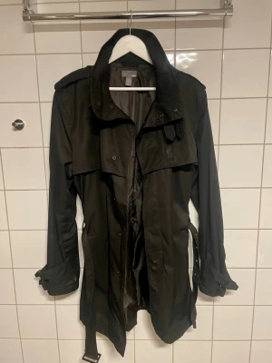 Svart trenchcoat med bälte - Stilren svart trenchcoat med klassisk krage och bälte i midjan. Spännen vid ärmsluten och ett slätt tyg som ger en snygg siluett. Perfekt för dig som gillar tidlös och ett cleant utseende 