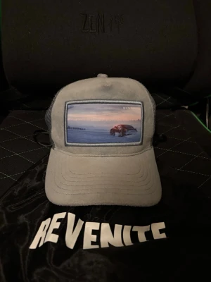 Grå keps från Revenite med bilmotiv - Snygg grå keps från Revenite med mesh på sidorna och ett coolt broderat motiv av en bil på framsidan. Kepsen har böjd skärm och justerbar snapback baktill. Perfekt för dig som gillar streetwear och unika accessoarer. OG keps