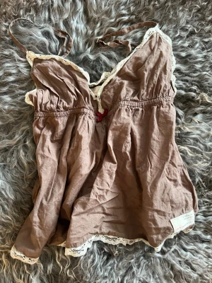 Brunt nattlinne med spetsdetaljer - Sött nattlinne i brun bomull med tunna axelband och dekorativ vit spets längs kanterna. Liten rosett framtill ger en gullig touch. Perfekt för mysiga kvällar hemma.