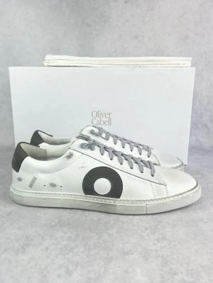 Vita sneakers från Oliver Cabell - Stilrena vita sneakers från Oliver Cabell med grå detaljer och grå skosnören. Skorna har en svart cirkeldetalj på sidan och svart hälparti. Tillverkade i läder med klassisk låg profil och vit sula. Perfekta för en clean och modern look.