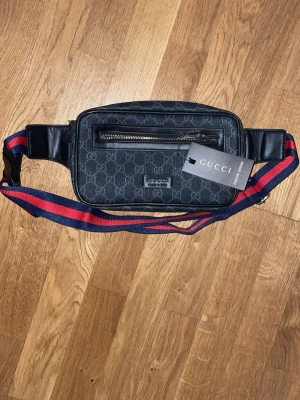 Svart midjeväska från Gucci - Snygg svart midjeväska från Gucci med klassiskt GG-mönster i grått. Väskan har en framficka med dragkedja och justerbar axelrem i rött och blått. Tillverkad i slitstarkt material med läderdetaljer och metallogo framtill. Perfekt för dig som vill ha en trendig och praktisk accessoar.