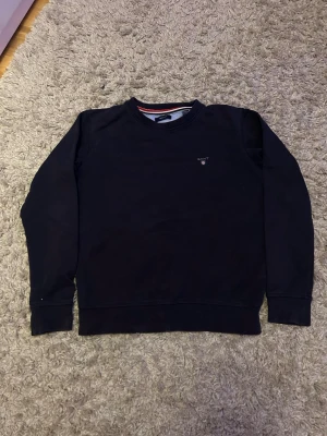 Mörkblå sweatshirt från Gant - En stilren mörkblå sweatshirt från Gant med rund halsringning och diskret logga på bröstet. Tröjan har långa ärmar och ribbade muddar vid ärmslut och nederkant. Perfekt för en avslappnad och snygg look. Med size 11-12 år 146-152
