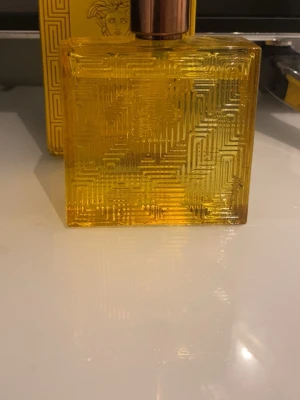 Versace gul parfymflaska - Snygg parfym från Versace i en rektangulär, gul glasflaska med geometriskt mönster. Flaskan har ett guldigt lock och den klassiska Versace-loggan syns på förpackningen. Perfekt för dig som gillar exklusiva dofter och stilren design.