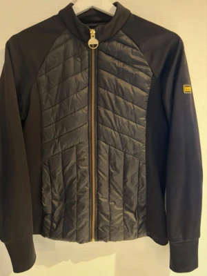 Barbour jacka - Svart Barbour International jacka med dragkedja framtill. Jackan har en quiltad front och slätare ärmar och rygg. Det finns två öppna fickor i sidorna. Barbour International-logotypen finns på vänster ärm och på dragkedjans handtag. Storlek 34, rökfritt hem