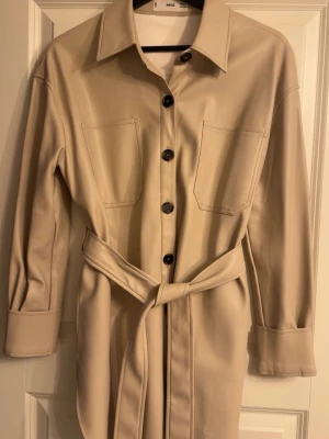 Beige skinnkappa med bälte från Mango - Beige skinnkappa från Mango i syntetmaterial med knappar framtill, två bröstfickor och matchande bälte i midjan. Kappan har klassisk krage och långa ärmar med breda manschetter.
