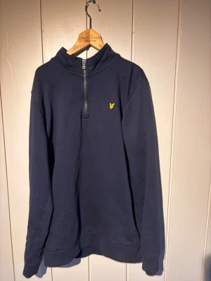 Mörkblå halfzip från Lyle & Scott - Mörkblå långärmad tröja från Lyle & Scott Junior med halv dragkedja och klassisk gul logga på bröstet. Tröjan har hög krage och är tillverkad i mjukt material, perfekt för en avslappnad stil.