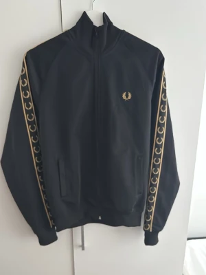 Svart Fred Perry taped track jacket - Cool svart track jacket från Fred Perry med guldiga detaljer och logga på bröstet. Guldiga band med Fred Perry-krans längs ärmarna, hög krage och dragkedja framtill. Perfekt för en sportig och stilren look. Använd 1 gång. Pris kan diskuteras vid snabb affär.