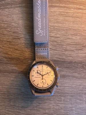 Omega x Swatch Speedmaster klocka - Säljer en stilren Omega x Swatch Speedmaster MoonSwatch med ljus urtavla och grått armband. Klockan har kronograf, svart bezel och tydliga index. Armbandet är i syntetmaterial med Speedmaster- och Swatch-logga. Perfekt för dig som gillar ikoniska samarbeten.
