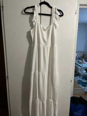 Vit långklänning med volangdetaljer - Lång, luftig vit klänning med breda axelband och volangdetaljer. Tillverkad i ett mjukt, strukturerat bomullstyg som ger en avslappnad och somrig känsla. Perfekt för dig som gillar enkel men stilren design.