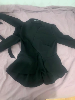 Svart kavaj med knytband från Zara - Stilren svart kavaj med markerad midja och knytband. Kavajen har en asymmetrisk skärning och långa ärmar. Perfekt för en trendig och modern look. Materialet känns mjukt och följsamt.