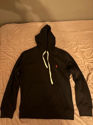 Svart hoodie från Polo Ralph Lauren - Snygg svart hoodie från Polo Ralph Lauren med klassisk huva och vita dragsnören. Har en röd broderad logga på bröstet och stor känguruficka framtill. Tillverkad i mjukt bomullsmaterial som är skönt mot huden. Perfekt för en chill och stilren look.