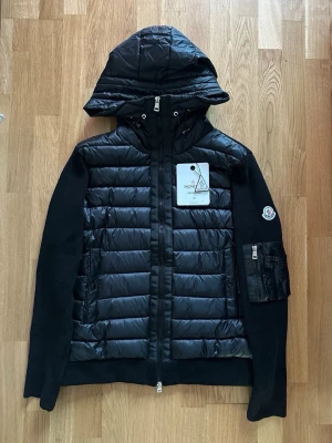 Säljer en moncler cardigan - Snygg svart dunjacka från Moncler med stickade ärmar och vadderad kropp. Jackan har en avtagbar huva, dragkedja framtill och fickor med dragkedja. Moncler-logga på ärmen och extra ficka med dragkedja på vänster ärm. Perfekt för kalla vinterdagar. Pris går att diskutera.