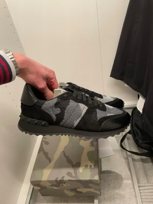 Valentino Garavani camo sneakers - Snygga sneakers från Valentino Garavani med svart och grått camomönster. Skorna har detaljer i mocka och läder, samt nitar på hälen. Tjock sula och klassisk snörning. Perfekta för dig som vill ha en unik och trendig look.