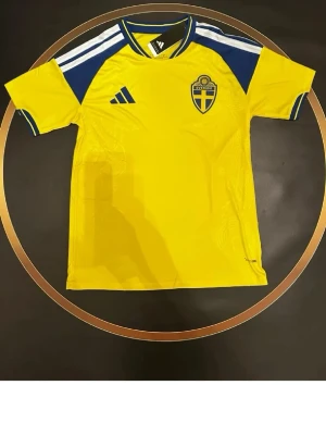 Svensk landslagströja Adidas gul/blå - Gul och blå svensk landslagströja från Adidas med broderad landslagssköld på bröstet och klassiska vita Adidas-ränder på axlarna. Tröjan har korta ärmar, rund hals och är tillverkad i ett lätt, ventilerande material perfekt för fotboll.