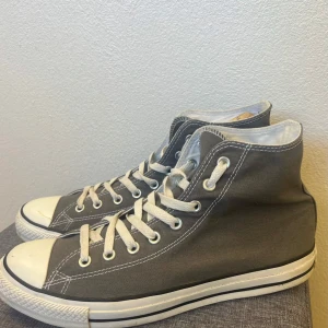 Grå höga Converse sneakers - Klassiska höga Converse sneakers i grå canvas med vita gummisulor och tåhättor. Skorna har vita snören, silverfärgade öljetter och kontrastsömmar. Perfekta för en avslappnad och cool streetstil. Storlek 44,5 