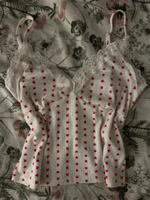 Vit body med röda hjärtan och spets - Söt vit body med tunna axelband och röda hjärtan över hela tyget.  Det står inte vilken storlek det är men jag tror att det är xs! V-ringad med vit spets längs halsringningen. Ribbad struktur och mjukt material som känns bekvämt mot huden. Perfekt för dig som gillar romantiska detaljer.