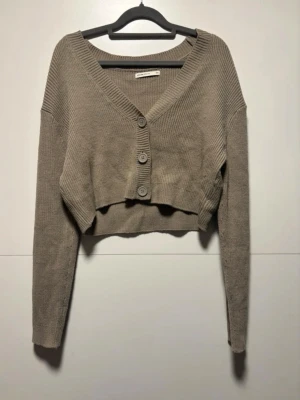 Beige cropped kofta med knappar - Trendig beige cropped kofta med tre stora knappar framtill och djup v-ringning. Långärmad och ribbstickad i mjukt material, perfekt att styla över ett linne eller t-shirt. Enkel och stilren design som passar till många outfits.