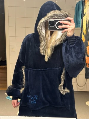 Snug Rug Eskimo Hoody - Snug Rug Eskimo Hoody! Storlek Onesize. Köpt för 695 kr på Roligaprylar. Helt oanvänd. Denna filt som är som en stor luvtröja är fodrad med supermjukt och varmt sherpafleece och har en ficka på magen.  Färg: MÖRKBLÅ Korsning mellan filt och fleece-hoodie Luva med sherpafleece på insidan och fuskpäls runt linningen av luvan Muddar längst ner på ärmarna  Stor fleecefodrad ficka på magen Hoodien är ca 97 cam lång i ryggen  Tvättas i maskin i 40 grader