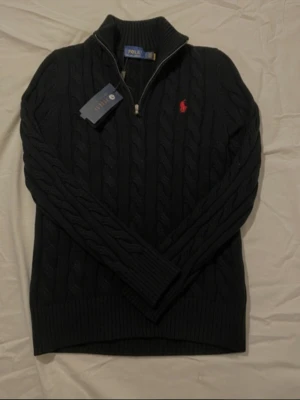 Mörkblå kabelstickad tröja Polo Ralph Lauren - Kabelstickad mörkblå tröja från Polo Ralph Lauren med halv dragkedja och röd broderad logga på bröstet. Tröjan har hög krage och ribbade muddar vid ärmslut och nederkant. Perfekt för dig som gillar klassisk och stilren design.