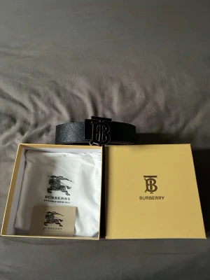 Svart skärp från BB med monogram - Snyggt svart skärp (inte äkta) men inspirerat från Burberry med präglat monogrammönster längs hela bandet och en stor metallspänne med deras ikoniska TB-logga. Går vända på, då är det mörkbrunt. Skärpet kommer i förpackning med dustbag och certifikat. Perfekt accessoar för att lyfta din outfit. 