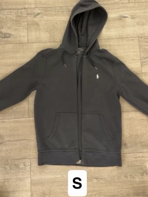 Svart hoodie från Polo Ralph Lauren - Snygg svart hoodie med dragkedja från Polo Ralph Lauren. Klassisk design med vit broderad logga på bröstet, två fickor fram och justerbar huva med snören. Perfekt för en chill och stilren look.