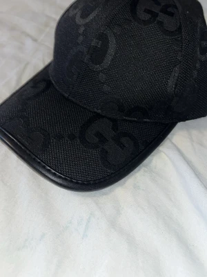 Svart keps med GG-mönster  - Snygg svart keps med det klassiska GG-monogrammet ton-i-ton över hela kepsen. Kepsen har böjd skärm med läderkant och är tillverkad i ett tåligt tygmaterial. Perfekt accessoar för en trendig och exklusiv look.