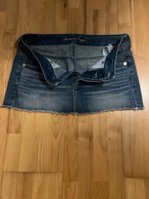 Blå jeanskjol med rå kant - Snygg blå jeanskjol i kort modell med rå, fransig kant nertill. Klassisk femficksdesign och slitna detaljer framtill. Perfekt för en avslappnad och trendig look.