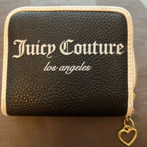 Juicy Couture svart plånbok - Snygg svart plånbok från Juicy Couture med vit kant och text. Plånboken har dragkedja med hjärtformat guldfärgat hänge, flera kortfack och ett fönsterfack. Materialet är konstläder och insidan är ljusbeige med gulddetaljer.