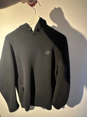 Svart Nike tech hoodie med huva - Säljer en svart hoodie från Nike med liten logga på bröstet. Hoodien har dragsko i huvan och en klassisk känguruficka framtill. Perfekt för en chill och sportig stil. Materialet känns mjukt och bekvämt.
