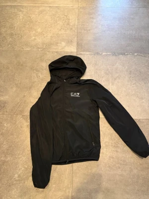 Svart EA7 vindjacka med huva - Säljer en svart vindjacka från EA7 Emporio Armani med huva och dragkedja framtill. Jackan har två fickor med dragkedja och logotypen tryckt på bröstet. Materialet är lätt och smidigt, perfekt för att skydda mot vind.