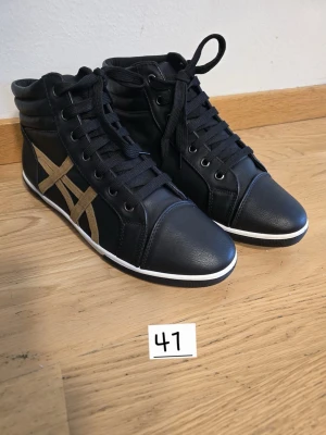 Svarta Asics high top sneakers - Snygga svarta high top sneakers från Asics med klassiska guldiga stripes på sidan. Skorna har svart snörning, vit sula och är tillverkade i ett slitstarkt syntetmaterial med en sportig vibe. Perfekta för dig som vill ha en clean och stilren look.