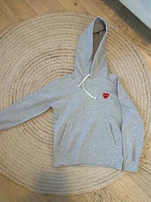 Grå hoodie Comme des Garçons Play - Säljer en grå hoodie från Comme des Garçons Play med det ikoniska röda hjärtat med ögon broderat på bröstet. Hoodien har huva med vit dragsko och två fickor framtill. Tillverkad i mjuk bomullsblandning, perfekt för en chill och stilren look.