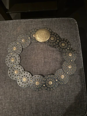 Svart bälte med runda metallplattor - Svart bälte med stora runda plattor i metall och dekorativa hål. Bältet har en snygg, antikguldig spänne med ingraverat mönster. Perfekt accessoar för att lyfta din outfit med en cool och annorlunda touch.