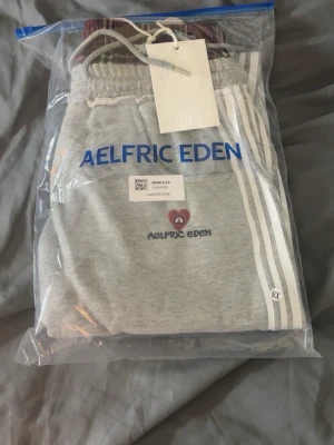 Grå mjukisbyxor från Aelfric Eden - Snygga grå mjukisbyxor från Aelfric Eden med vita ränder längs sidorna och ett litet rött hjärta broderat framtill. Byxorna har dragsko i midjan och är gjorda i mjukt material, perfekta för chill dagar. Stilen är avslappnad och trendig.