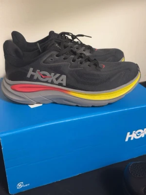 Hoka svarta sneakers med färgad sula - Säljer ett par svarta Hoka sneakers med grå, gul och röd detaljer på sulan. Skorna har en låg profil och är tillverkade i mesh-material med klassiska snörningar. Perfekta för dig som gillar sportig och modern stil.