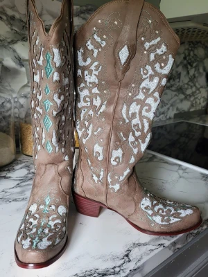Cowboy Boots stl. 35 - Snygga beige cowboy boots med högt skaft och spetsig tå. Amerikanskt mönster i vitt och turkost med strasstenar och glitter som ger en unik look.                                      Perfekta för dig som vill sticka ut.                         Innermått: 22,5 cm