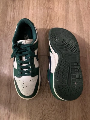 Nike Dunk Low grön/vit sneaker - Nike Dunk Low sneakers i grönt och vitt med mörkblå Swoosh. Skorna har klassisk låg siluett, perforerad tåbox och gröna skosnören. Ovandelen är i läder och yttersulan är i gummi för bra grepp. Perfekt för dig som gillar retrostil och streetwear. Äkta