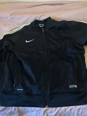 Svart Nike Dri-FIT träningsjacka - Snygg svart träningsjacka från Nike med vit logga på bröstet och vita detaljer på axlarna. Jackan har hel dragkedja, två sidofickor och är tillverkad i Dri-FIT-material som transporterar bort fukt. Perfekt för träning eller uppvärmning.