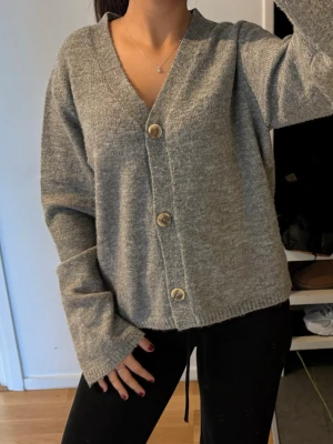 Grå kofta med träknappar - Mysig grå kofta med v-ringning och stora träknappar framtill. Koftan har en avslappnad passform, långa ärmar och ribbade muddar. Perfekt att slänga på sig över en t-shirt eller linne för en chill look.