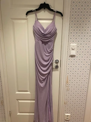 Ljuslila glittrig långklänning storlek S - Säljer en oanvänd ljuslila långklänning köpt i Maiami med smala axelband och draperad front. Klänningen har glittriga detaljer över hela tyget och en figurnära passform med slits framtill. Perfekt för bal eller för större evenemang!