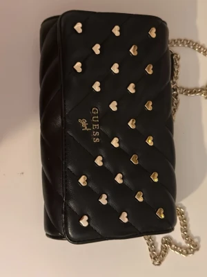 Svart quiltad handväska från Guess - Snygg svart quiltad handväska från Guess med guldfärgade och silverfärgade hjärtformade nitar på framsidan. Väskan har kedjerem i metall och ett ljust innerfoder med logomönster. Perfekt för dig som gillar detaljer och trendig design.