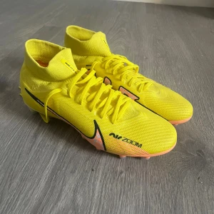 Nike Mercurial Zoom fotbollsskor gul Pros - Säljer ett par Nike Mercurial Zoom fotbollsskor i en grymt snygg gul färg med svart och orange detaljer. Skorna har hög krage, snörning och en lätt, ventilerande ovandel i syntetmaterial. Sulan har fasta dobbar för spel på naturgräs. Perfekt för dig som vill ha snabbhet och kontroll på planen.